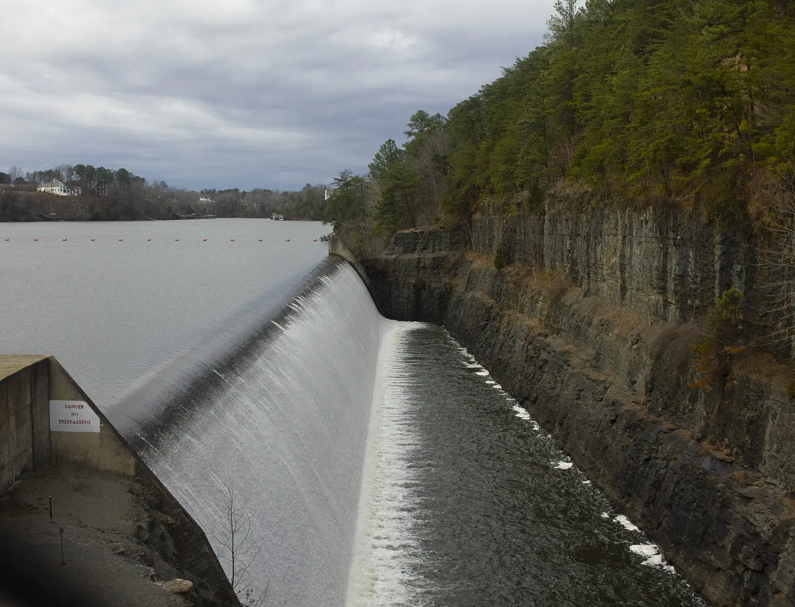 Water spills over the Lake Tuscaloosa dam spillway, with homes on the lake shore beyond.